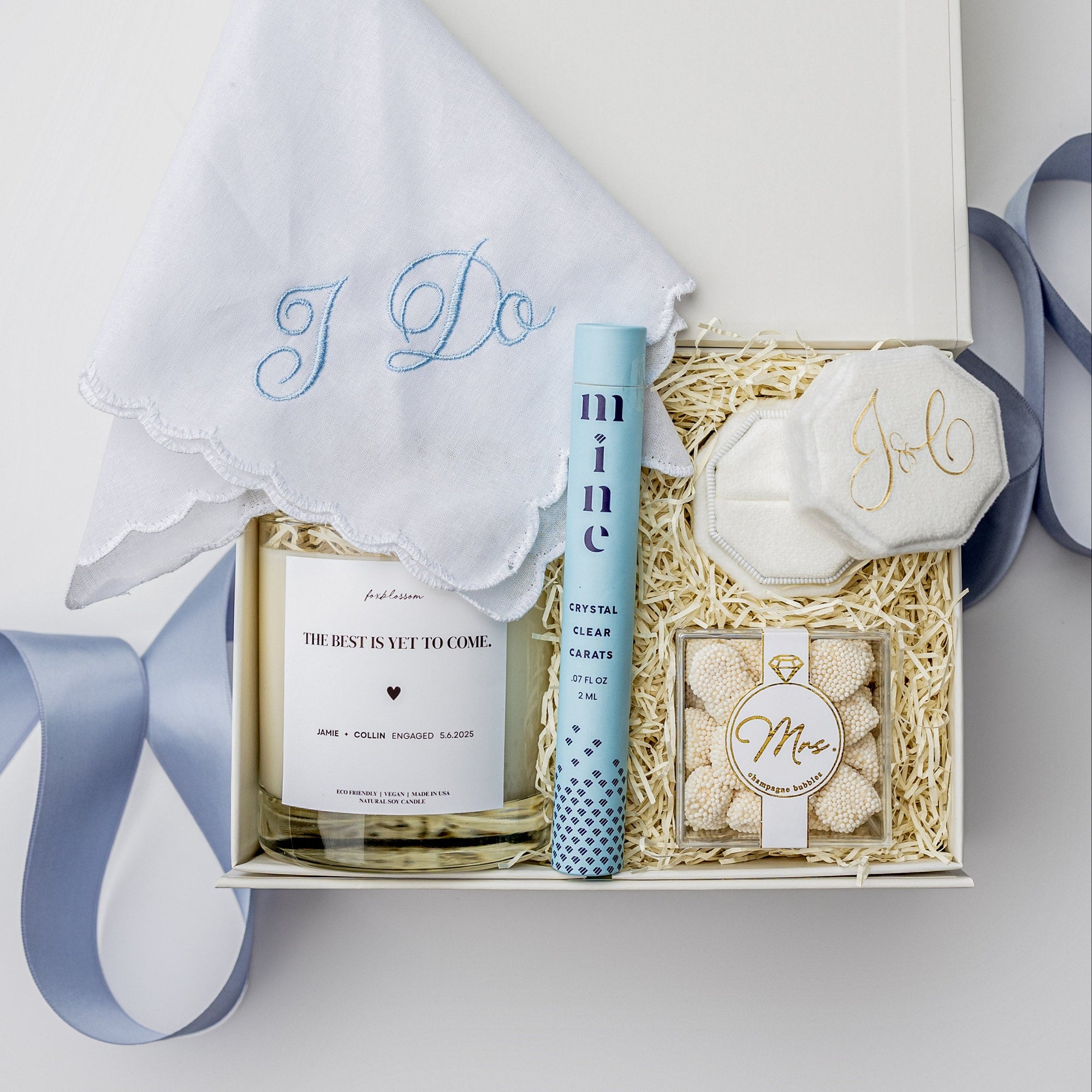 Top view of a “Something Blue” bride to be engagement gift box displayed in a white box with a blue ribbon and ivory shred, containing an embroidered “I Do” handkerchief, a ring cleansing pen, a personalized velvet ring box, an 8oz “Bride’s Bouquet” personalized candle, and Mrs. Champagne Bubbles gummies.