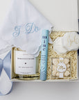 Top view of a “Something Blue” bride to be engagement gift box displayed in a white box with a blue ribbon and ivory shred, containing an embroidered “I Do” handkerchief, a ring cleansing pen, a personalized velvet ring box, an 8oz “Bride’s Bouquet” personalized candle, and Mrs. Champagne Bubbles gummies.