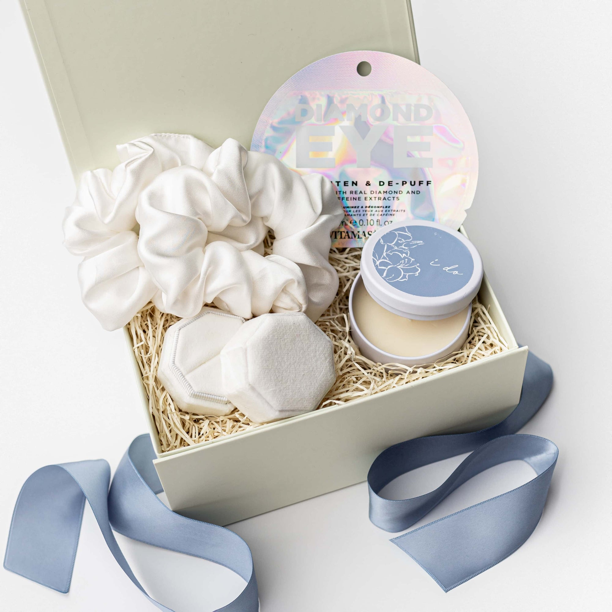 Engagement Gift Box for the Bride to Be, A top view of the Something Blue Gift Box presented in a white gift box filled with ivory shred and tied with a blue ribbon, featuring a set of two white silk scrunchies, a white velvet ring box with optional personalization, a diamond eye mask, and a 4 oz. hand-poured “I Do” candle, all arranged on a white background.