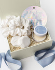 Engagement Gift Box for the Bride to Be, A top view of the Something Blue Gift Box presented in a white gift box filled with ivory shred and tied with a blue ribbon, featuring a set of two white silk scrunchies, a white velvet ring box with optional personalization, a diamond eye mask, and a 4 oz. hand-poured “I Do” candle, all arranged on a white background.