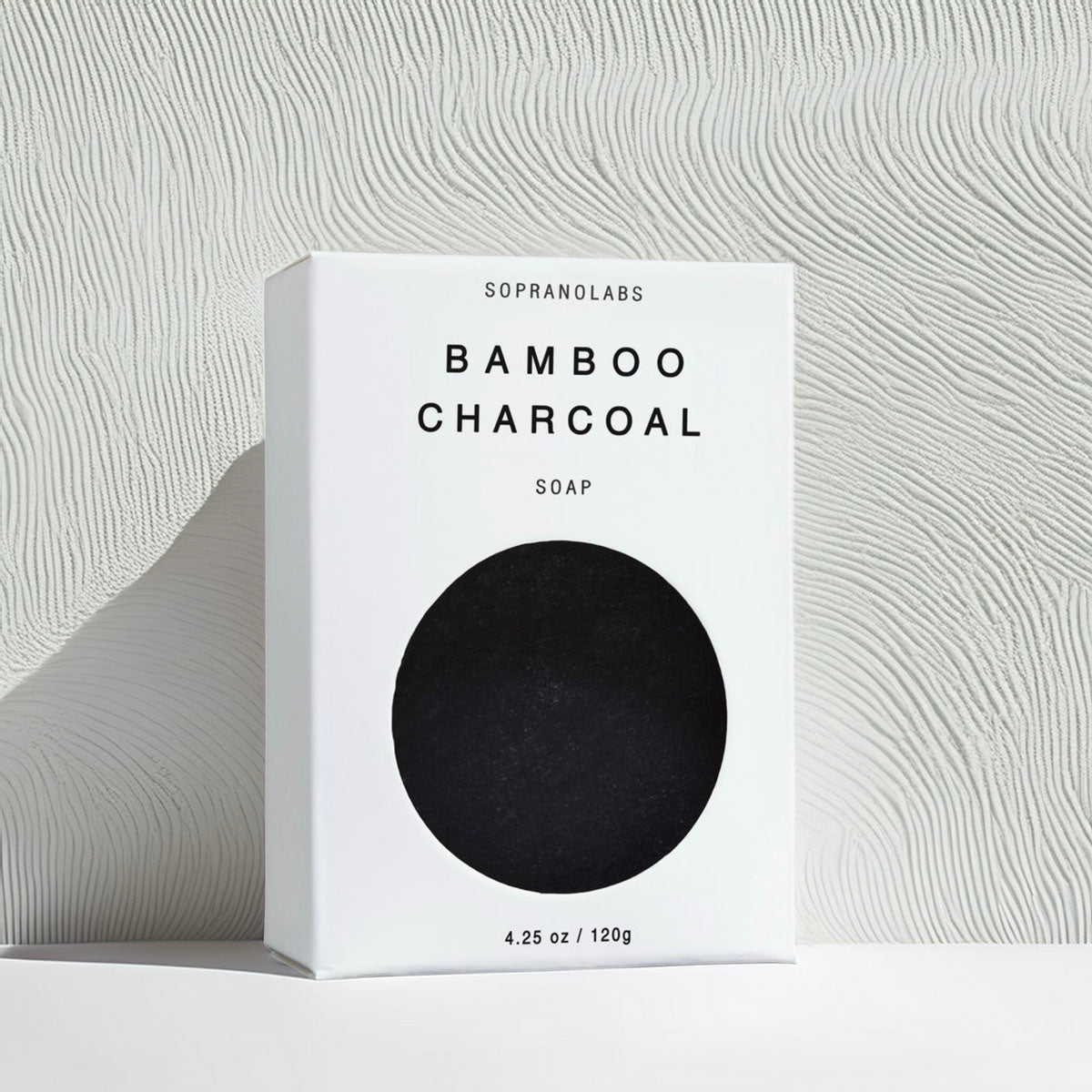 A white rectangular box of Sopranolabs Bamboo Charcoal Soap stands on a white surface against a textured light gray wall. The front of the box has a large black oval cutout and black text reading 'SOPRANOLABS BAMBOO CHARCOAL SOAP' and '4.