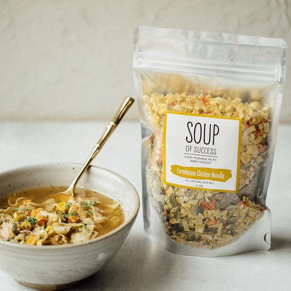 Soup of success, farmhouse chicken noodle, all natural soup mix in its front transparent with grey back rectangular foil container with a bowl of chicken soup next to it with a gold spoon in a white background. 