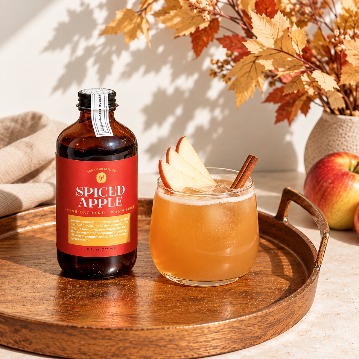 A bottle of Spiced Apple flavored non-alcoholic malt beverage sits on a round wooden tray next to a glass of the amber-colored drink, garnished with apple slices and a cinnamon stick, with apples and autumn leaves in the background.