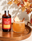 A bottle of Spiced Apple flavored non-alcoholic malt beverage sits on a round wooden tray next to a glass of the amber-colored drink, garnished with apple slices and a cinnamon stick, with apples and autumn leaves in the background.
