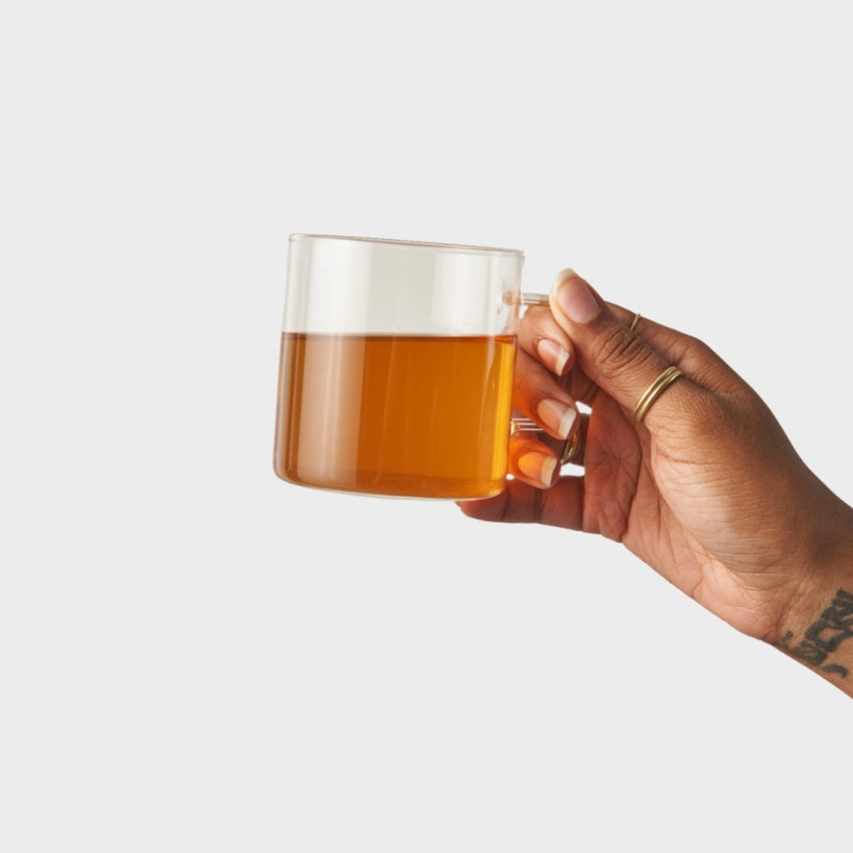 A hand holding a clear glass mug filled with amber tea against a plain light gray background.