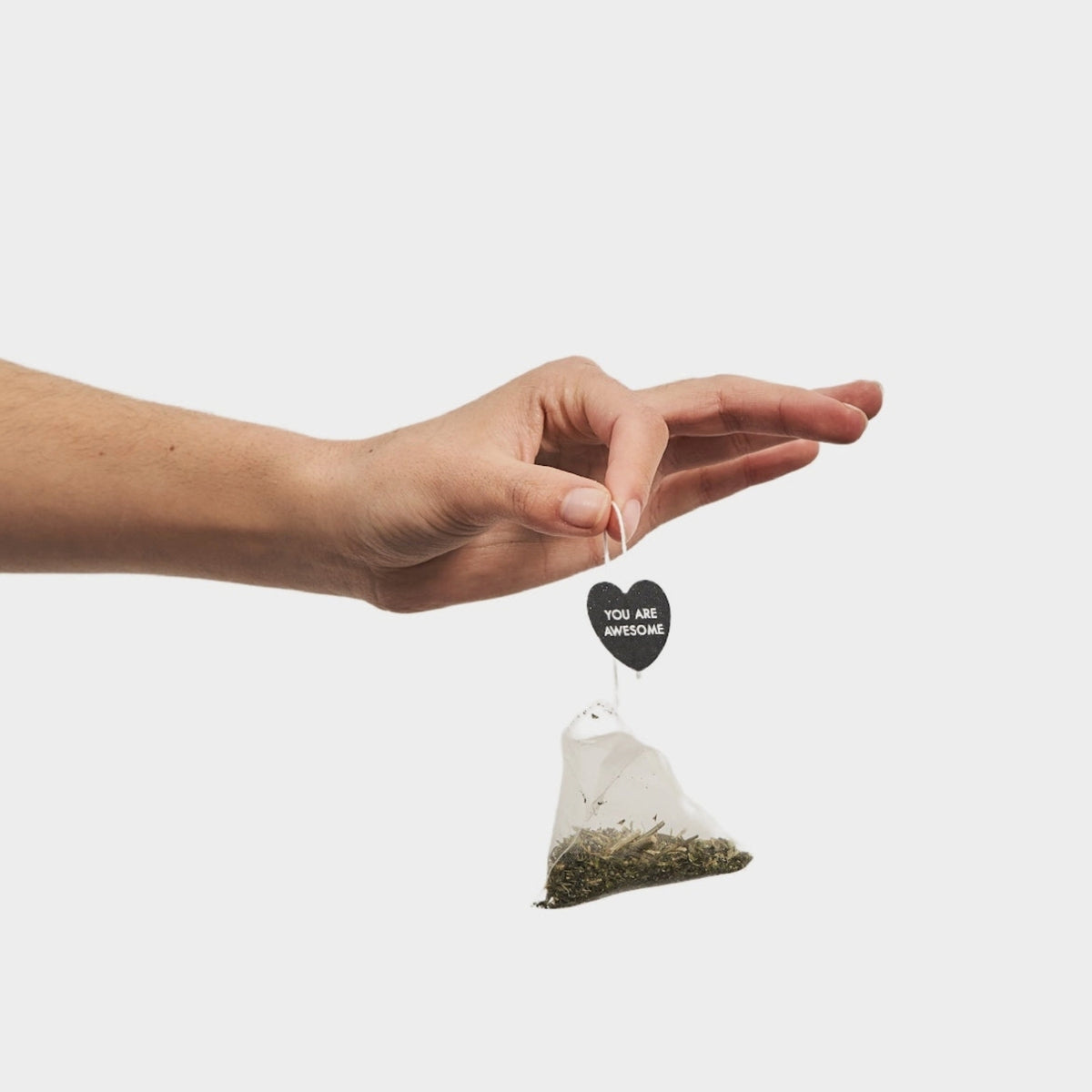 A hand holding a triangular tea bag charm with a small black heart tag that reads 'YOU ARE WELCOME'. The tea bag contains loose green tea leaves and is photographed against a solid light gray background.