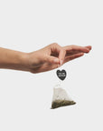 A hand holding a triangular tea bag charm with a small black heart tag that reads 'YOU ARE WELCOME'. The tea bag contains loose green tea leaves and is photographed against a solid light gray background.