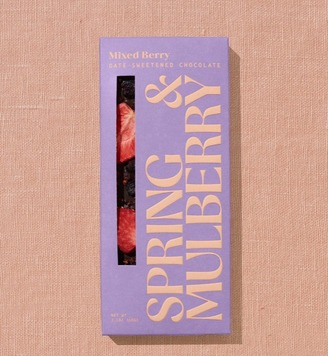Spring & Mulberry mixed berries chocolate bar in its rectangular package in Lila color with yellow lettering and a transparent part showing a little of the chocolate that is dark with pieces of strawberry on a beige background. 