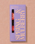 Spring & Mulberry mixed berries chocolate bar in its rectangular package in Lila color with yellow lettering and a transparent part showing a little of the chocolate that is dark with pieces of strawberry on a beige background. 