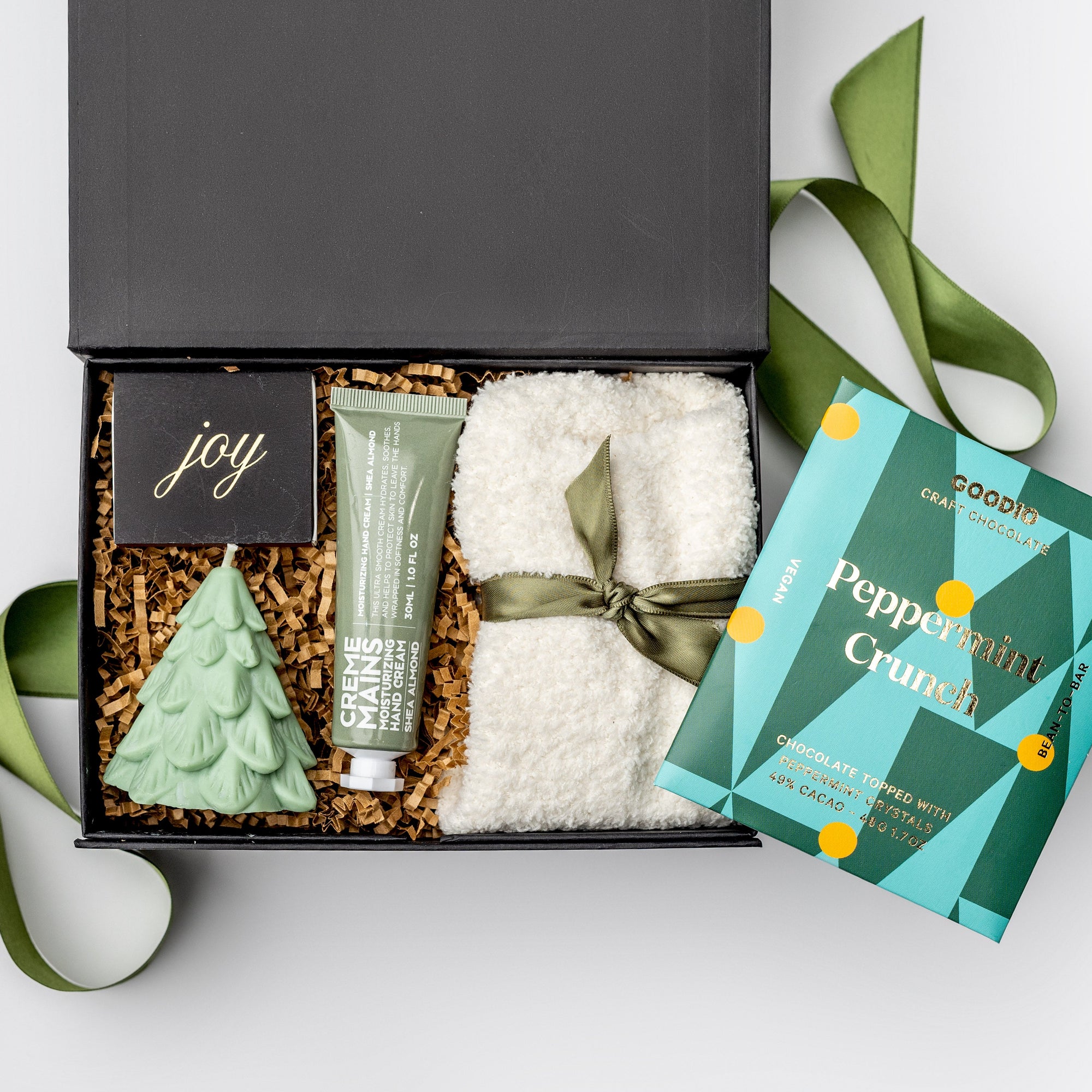 A black gift box filled with brown paper shred and tied with a green ribbon on a white background. Inside the box are unisex cozy cloud socks, an almond shea butter hand lotion, a festive foil-stamped matchbox, a hand-poured “Spruce Tree” scented candle, and a Goodio artisan peppermint crunch chocolate bar — a “Spruce and Joy” holiday gift box.
