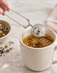 A stainless steel tea infuser held over a speckled cream ceramic mug filled with amber tea and loose flower petals. A hand is visible gripping the handle, with a small bowl of loose tea and a folded cloth on a light marble surface in the background.