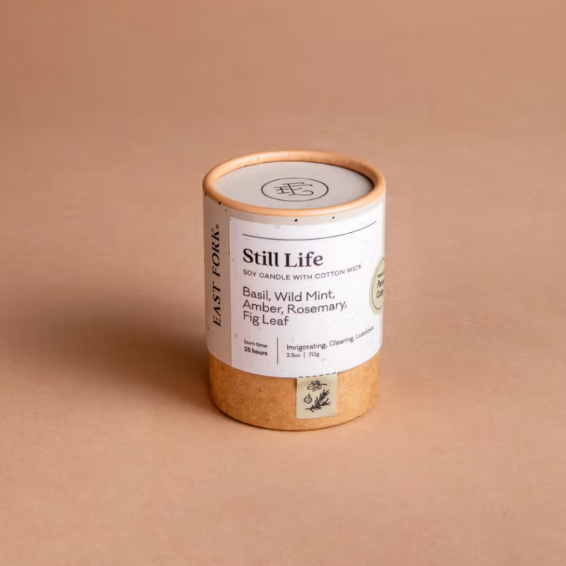 Still life candle is featured in its cylindrical container with tan bottom and rim white label black lettering on a beige background. 