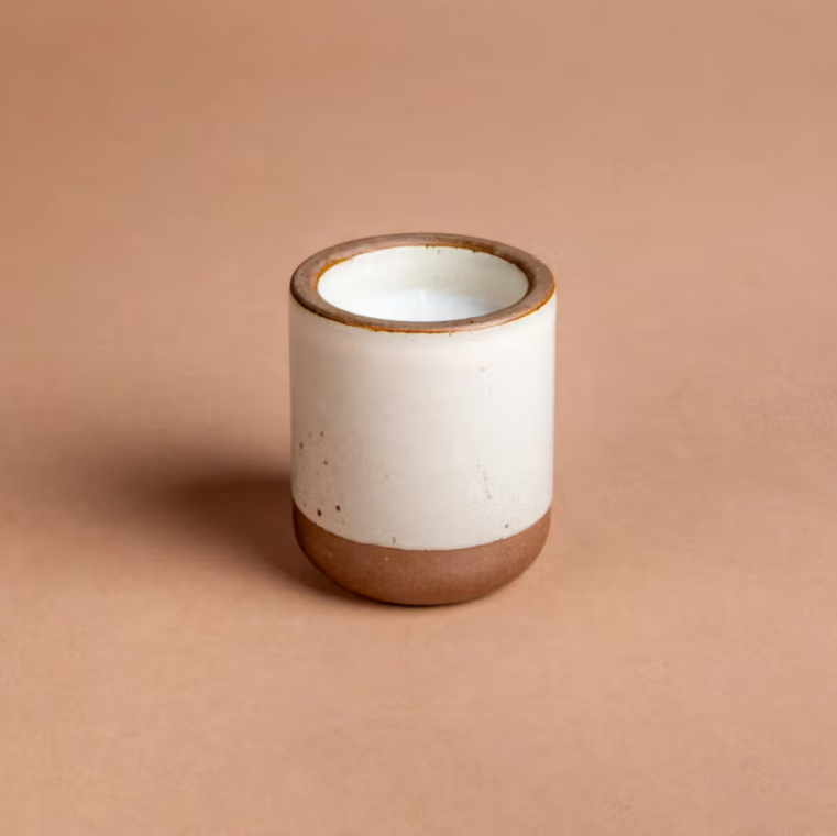 Still life candle is featured in its white ceramic jar with tan bottom and rim in a beige background. 