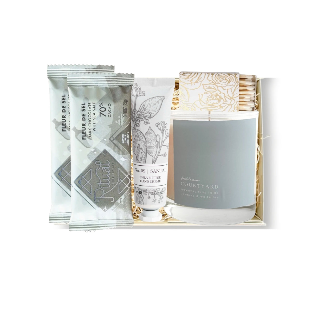 A curated gift set in a white box containing a gray candle, two silver chocolate bars, a white hand cream tube, and a pack of cotton pads, photographed on a white background.