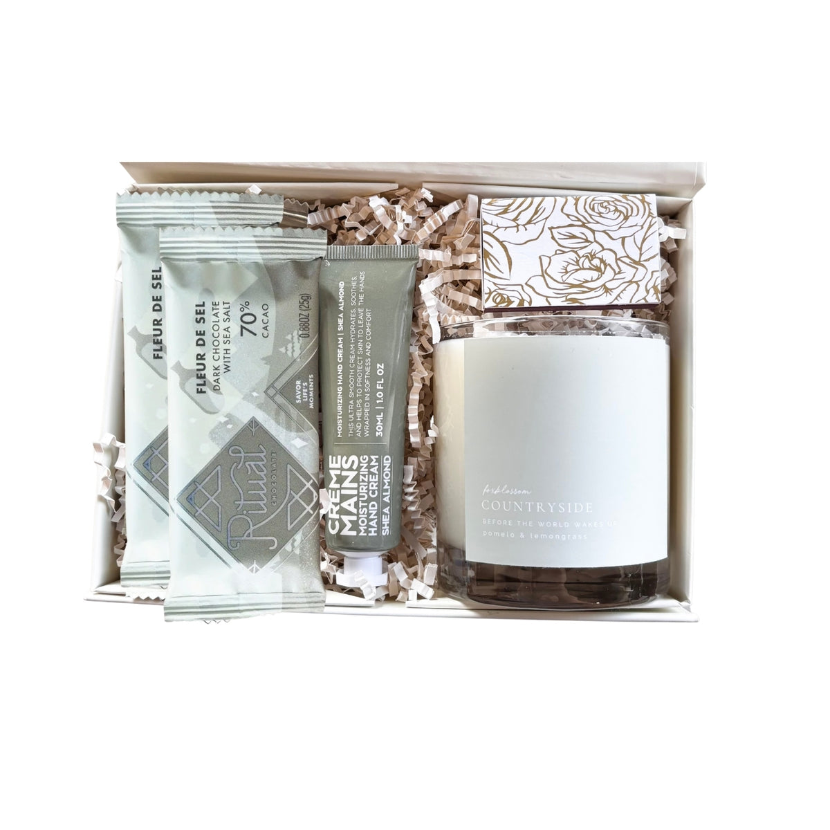 A curated self-care gift set in a white box with shredded paper filler, featuring a jar of 'Countryside' scented body oil, a tube of 'Creme' hand cream, two packets of 'Eau de Gel' hand gel, and a decorative box of bath salts.