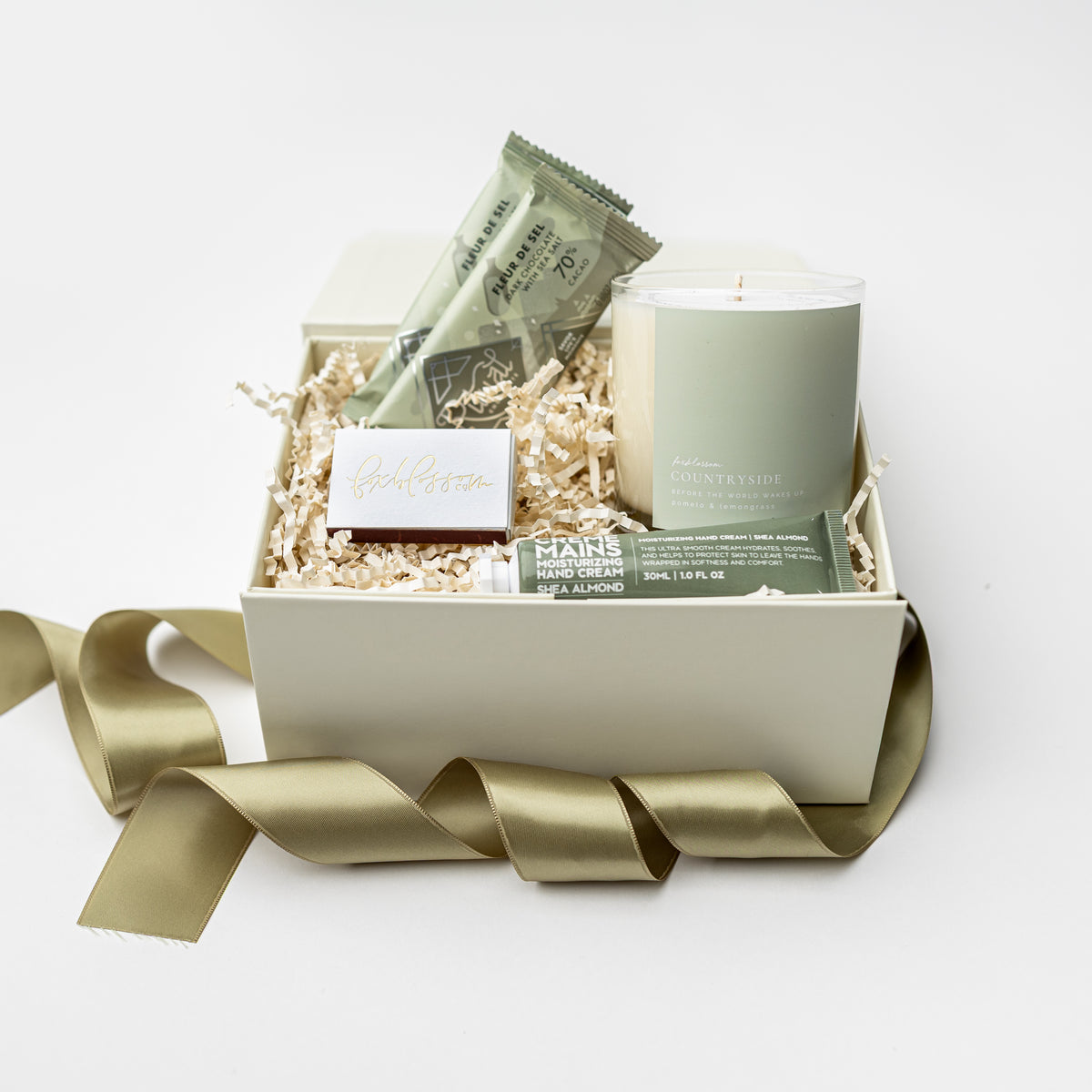 A curated gift box set on a white background, featuring a sage green candle, a chocolate bar, a small soap, and a bath towel arranged in white crinkle paper inside a cream-colored box with a gold satin ribbon.
