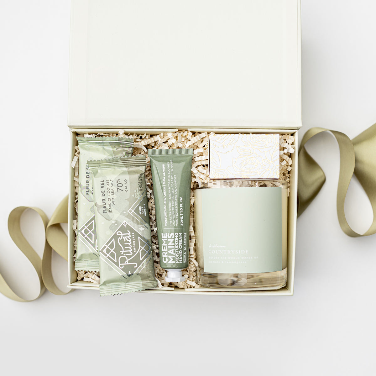 An open gift box containing a pale green candle, a tube of hand cream, two wrapped soap bars, and a small box, arranged in shredded paper with a ribbon around it on a white background.
