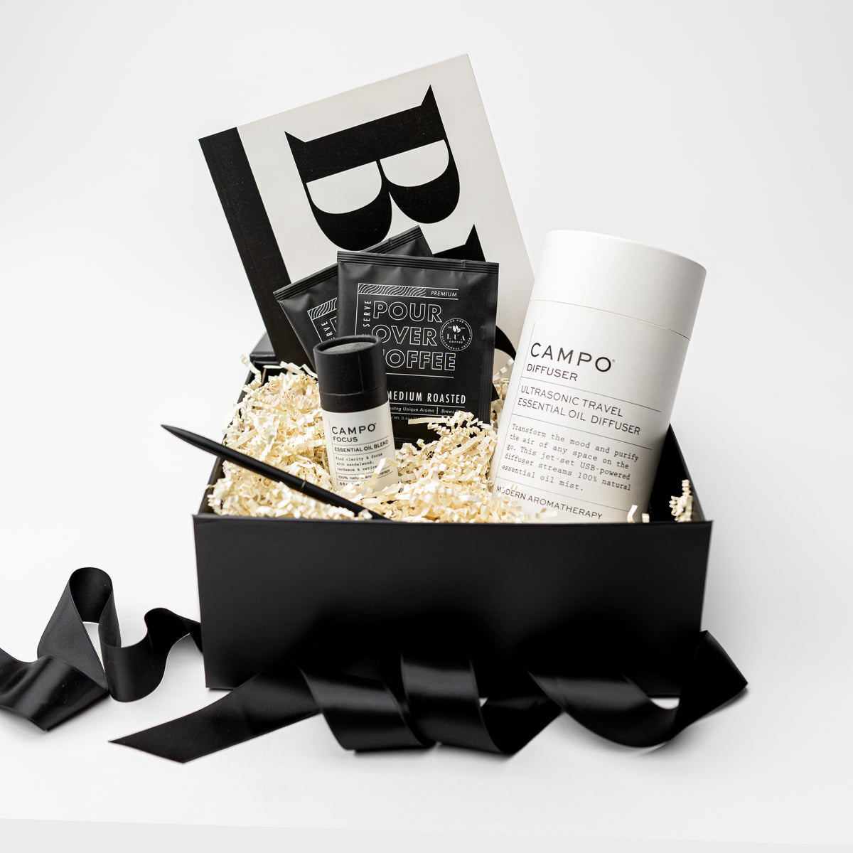A black gift box filled with spa and self-care items, including a white and black CAMPO diffuser, a small black bottle, two black CAMPO Pour Over Coffee sachets, and a black pen, arranged in white crinkle paper with a black ribbon in front on a white background.