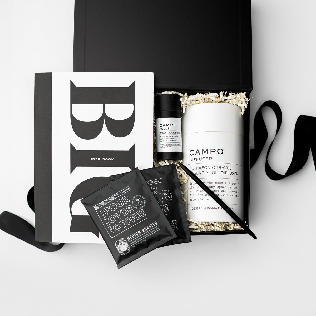 A curated gift set in a black box featuring a white and black Idea Book, a white CAMPO diffuser, two black COOFIE medium roasted coffee sachets, and a black pen, arranged on a light gray background.