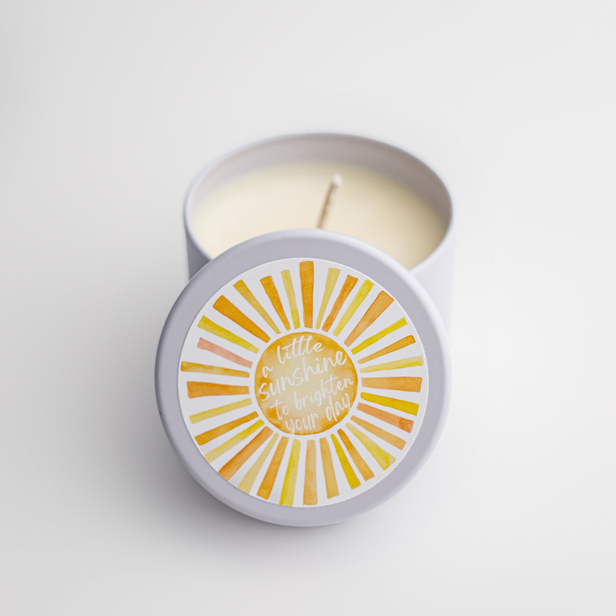 Sunshine natural soy candle in its white ceramic jar and white round label on top of the lid with a sun and in white lettering in the middle of it a little sunshine to brighten your day on a white background. 
