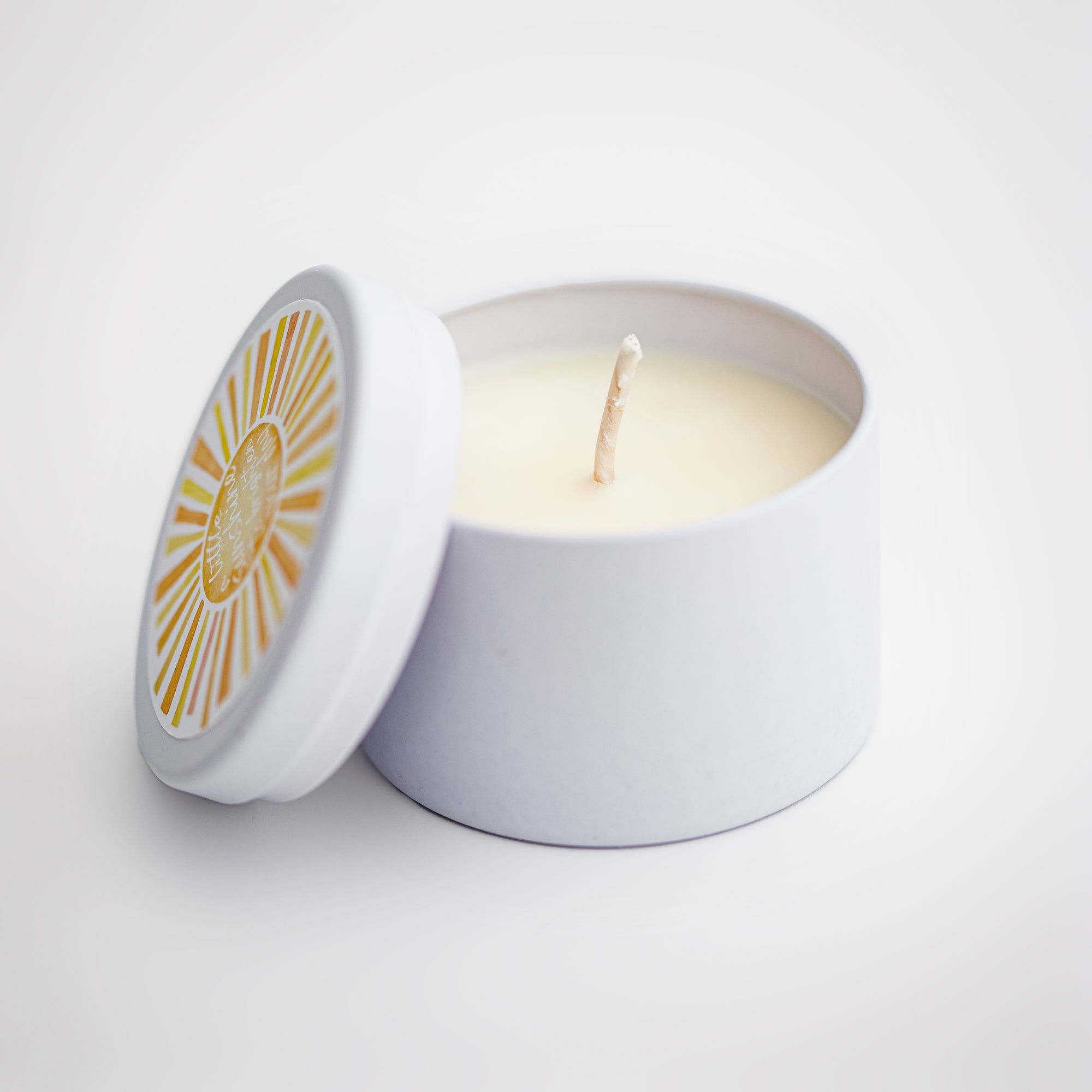 Sunshine natural soy candle in its white ceramic jar and white round label on top of the lid with a sun and in white lettering in the middle of it a little sunshine to brighten your day on a white background. 