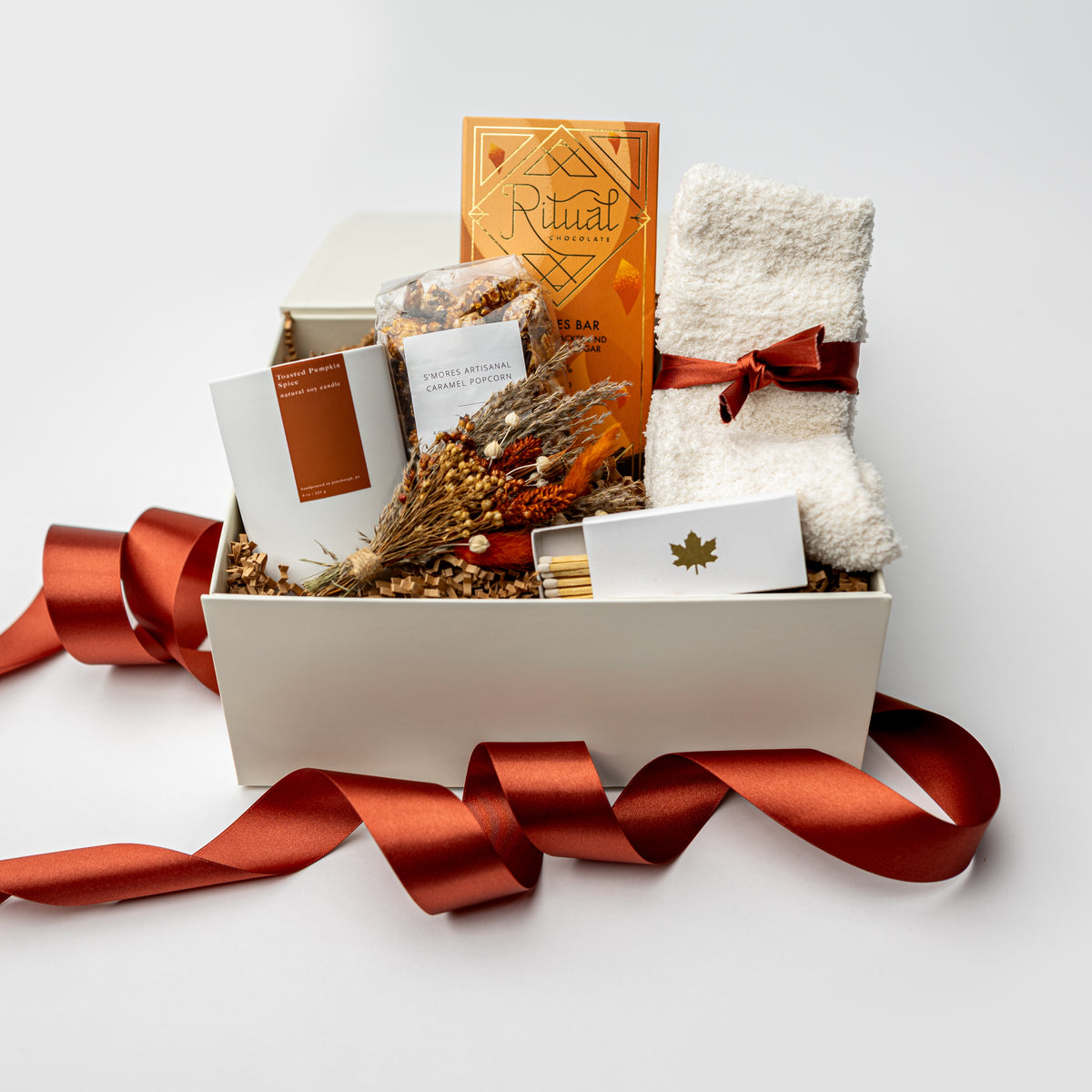 A curated gift box set on a white background, featuring a white box filled with dried botanicals, a white towel tied with a red ribbon, a Ritual candle, a white bath bomb package, and a matchbox, with a red satin ribbon draped in front.