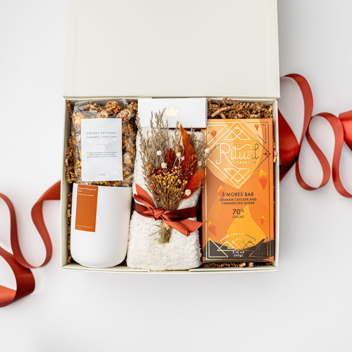 An open gift box arranged in a flat lay on a white background with red ribbon accents. The box contains a white candle, a white mug with an orange label, a small bundle of dried botanicals tied with a red ribbon, and an orange Ritual 3 Mores Bar package.