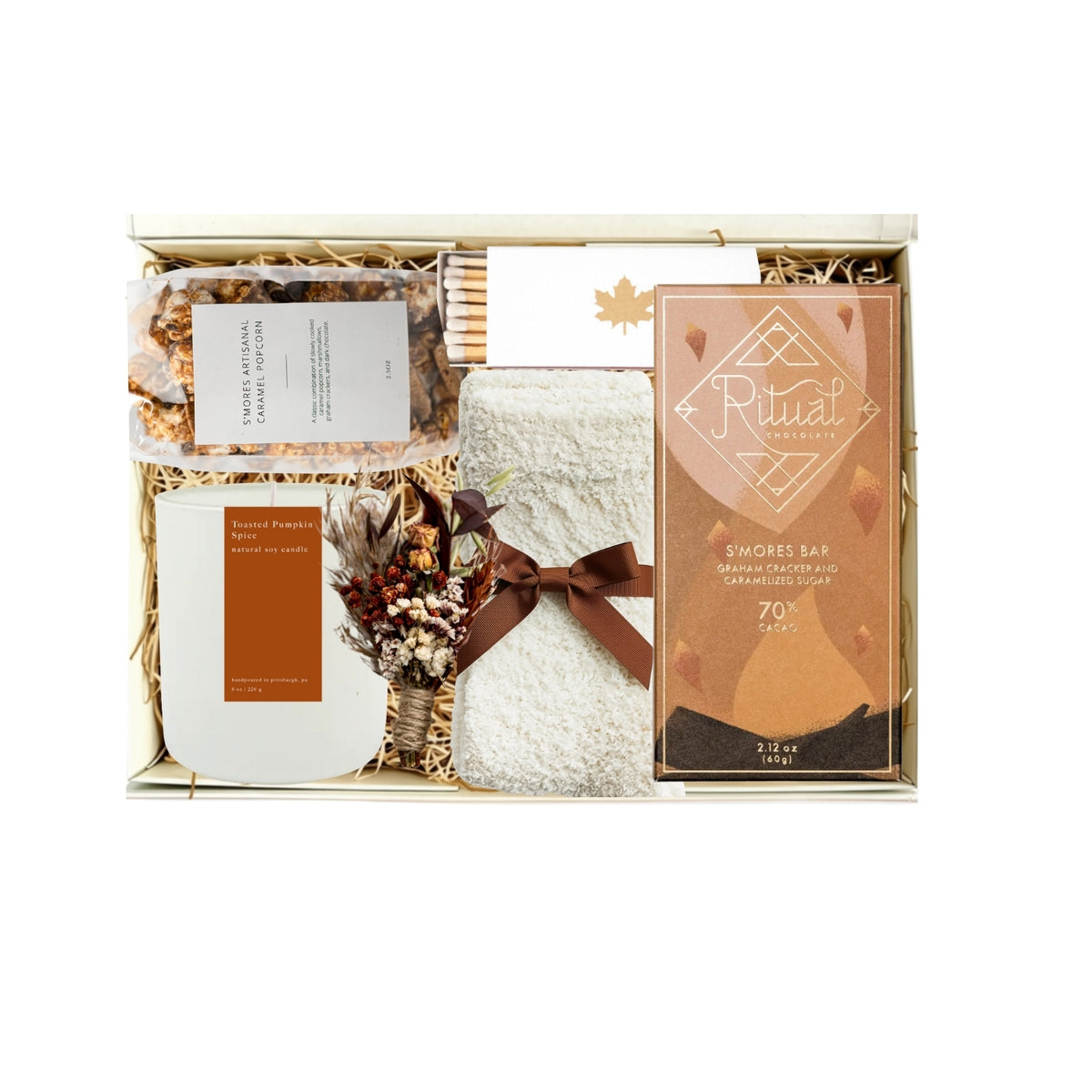 A curated gift box set containing a Ritwal chocolate bar, a white candle, a bag of coffee beans, and a small dried floral arrangement in a beige gift box with shredded paper.