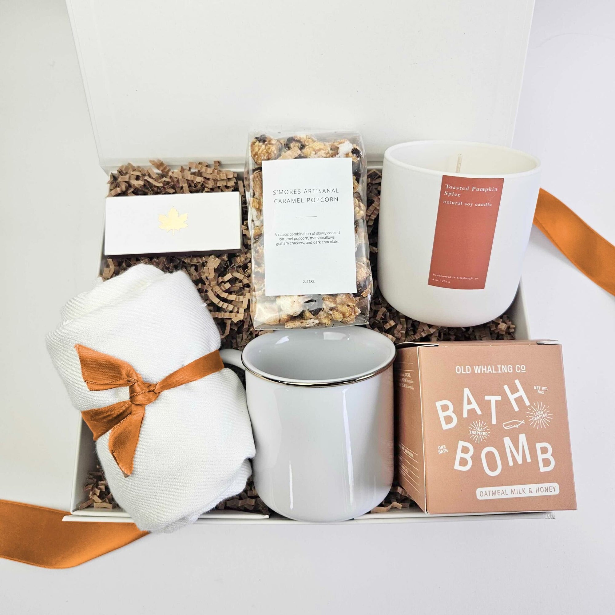 A gift box filled with brown paper shred and tied with an orange ribbon on a white background. Inside the box are a soft pashmina, s’mores popcorn, a maple-themed matchbox, a 9oz pumpkin spice candle, an oatmeal and honey bath bomb, and a gold-rim ceramic camp mug — a warm autumn-inspired gift set.