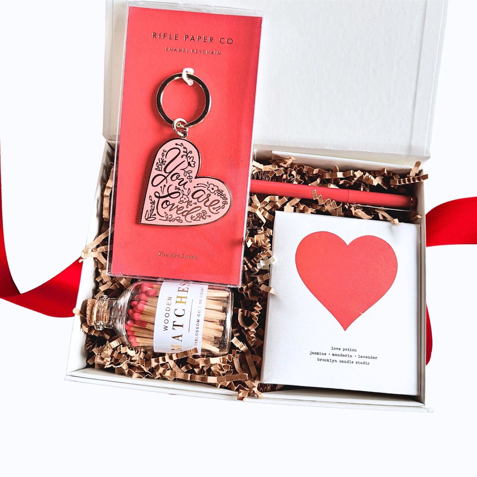 Open white gift box with a red ribbon filled with kraft crinkle paper, containing a heart-shaped keychain on red backing card, a red pen, a small bottle of wooden matches with red tips, and the love potion candle in a white box with a red heart printed on the lid.