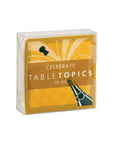 A box of cards, featuring a champagne bottle design, is presented against a plain white background. The box is a clear plastic covering a card with a design featuring a champagne bottle with a cork popping out, surrounded by celebratory dots and lines.  The words "CELEBRATE", "TABLETOPICS", and "TO GO" are printed on the card, in a bold and stylized font
