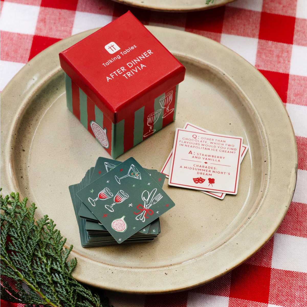 A boxed card game called Talking Tossers After Dinner Trivia sits on a beige ceramic plate over a red-and-white checkered tablecloth. The red box has green stripes and a small illustration.