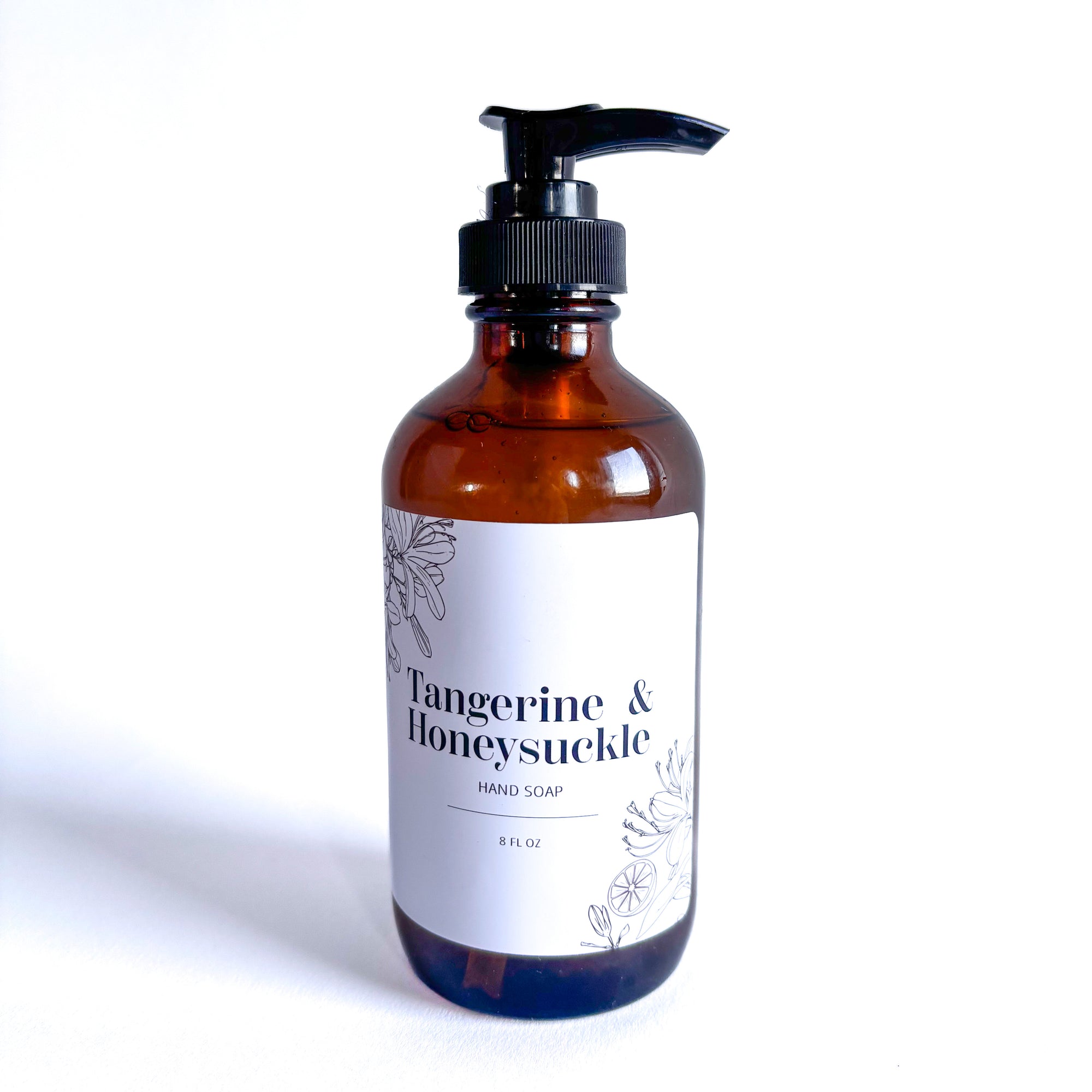 Tangerine and honeysuckle hand soap  in its cylindrical amber container and black pump is featured with white label with black lettering and flower with tangerine black silhouette on a white background.