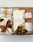 A top view of the Tea & Chocolate Gift Box presented in a white gift box filled with ivory shred and tied with a brown ribbon, featuring a 10 oz. speckled ceramic mug, ivory plush cloud socks, four salted caramel shortbread cookies, a crunchy caramel artisanal chocolate bar, two rooibos tea sachets, a gold stir spoon, and a handmade dried floral mini bouquet, all arranged on a white background.