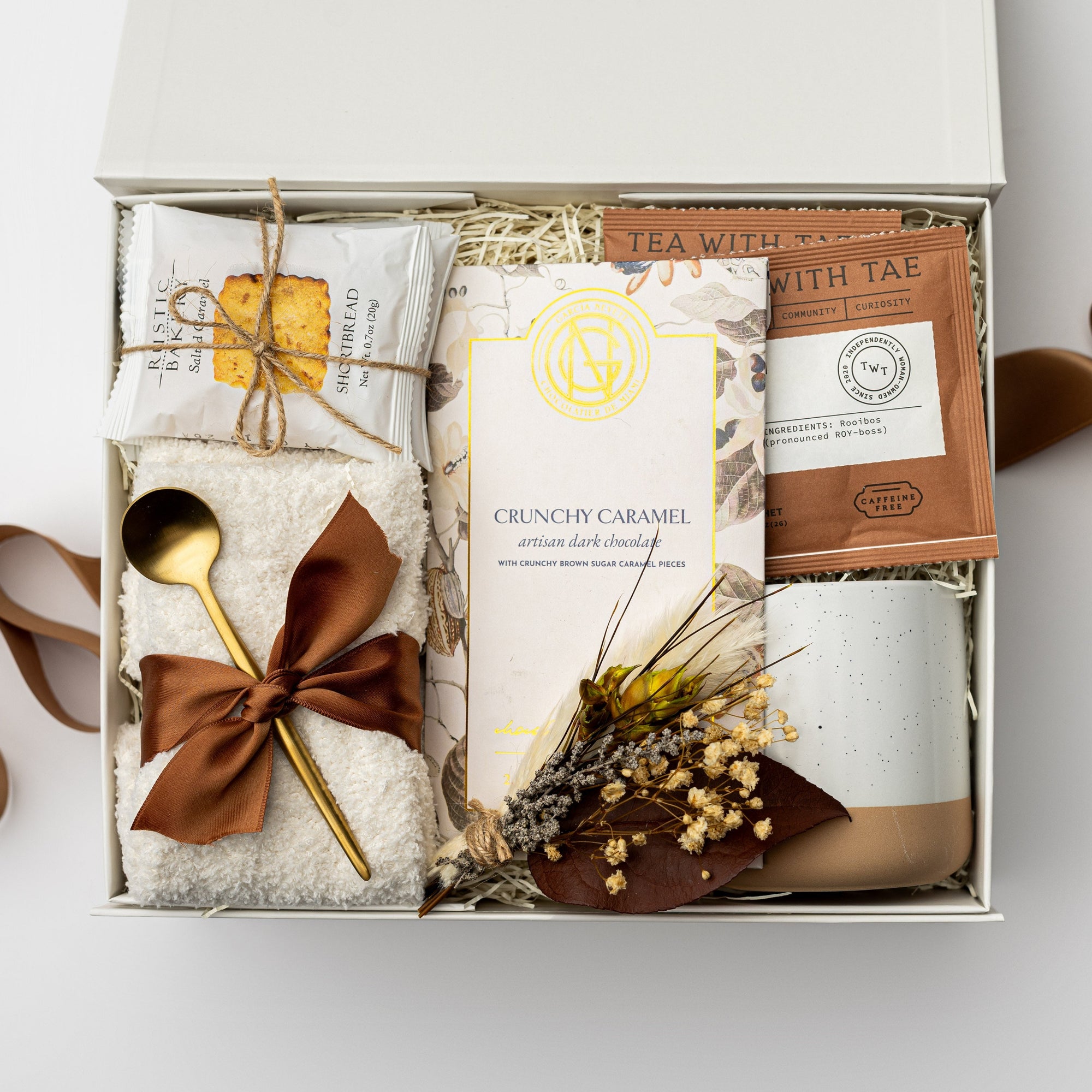 A top view of the Tea & Chocolate Gift Box presented in a white gift box filled with ivory shred and tied with a brown ribbon, featuring a 10 oz. speckled ceramic mug, ivory plush cloud socks, four salted caramel shortbread cookies, a crunchy caramel artisanal chocolate bar, two rooibos tea sachets, a gold stir spoon, and a handmade dried floral mini bouquet, all arranged on a white background.