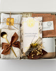 A top view of the Tea & Chocolate Gift Box presented in a white gift box filled with ivory shred and tied with a brown ribbon, featuring a 10 oz. speckled ceramic mug, ivory plush cloud socks, four salted caramel shortbread cookies, a crunchy caramel artisanal chocolate bar, two rooibos tea sachets, a gold stir spoon, and a handmade dried floral mini bouquet, all arranged on a white background.
