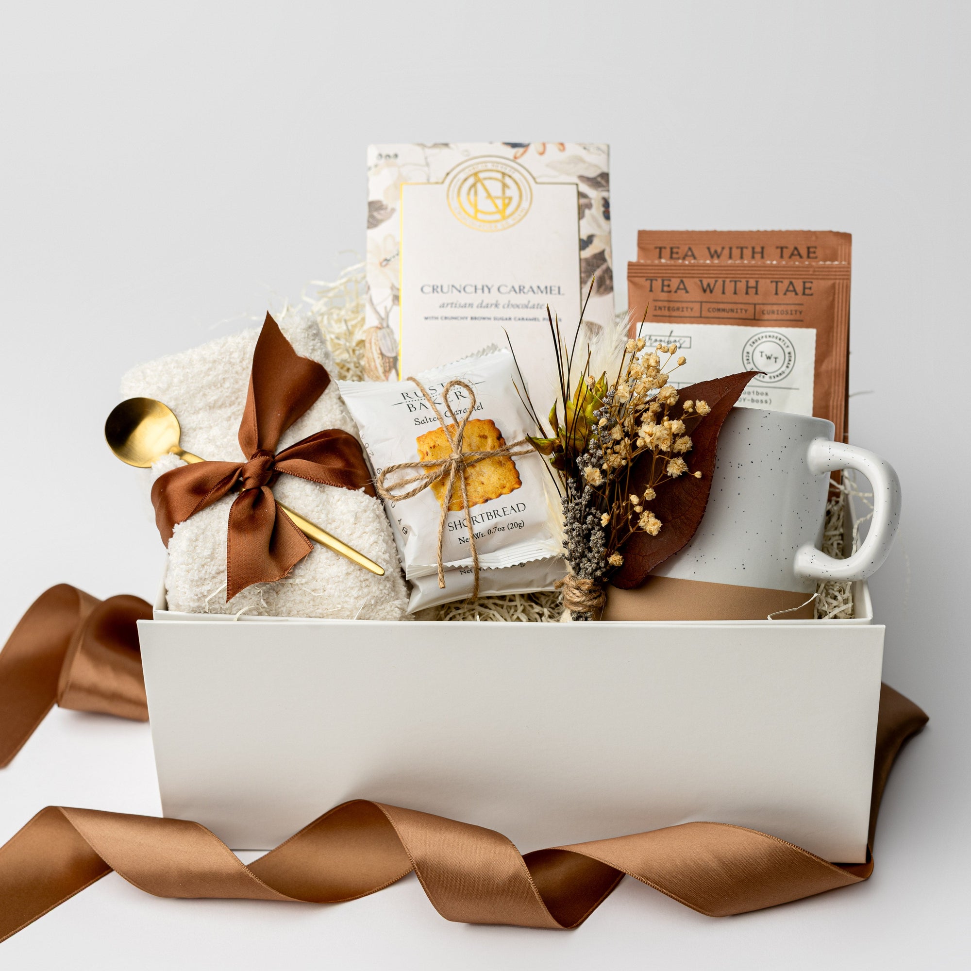 A top view of the Tea & Chocolate Gift Box presented in a white gift box filled with ivory shred and tied with a brown ribbon, featuring a 10 oz. speckled ceramic mug, ivory plush cloud socks, four salted caramel shortbread cookies, a crunchy caramel artisanal chocolate bar, two rooibos tea sachets, a gold stir spoon, and a handmade dried floral mini bouquet, all arranged on a white background.