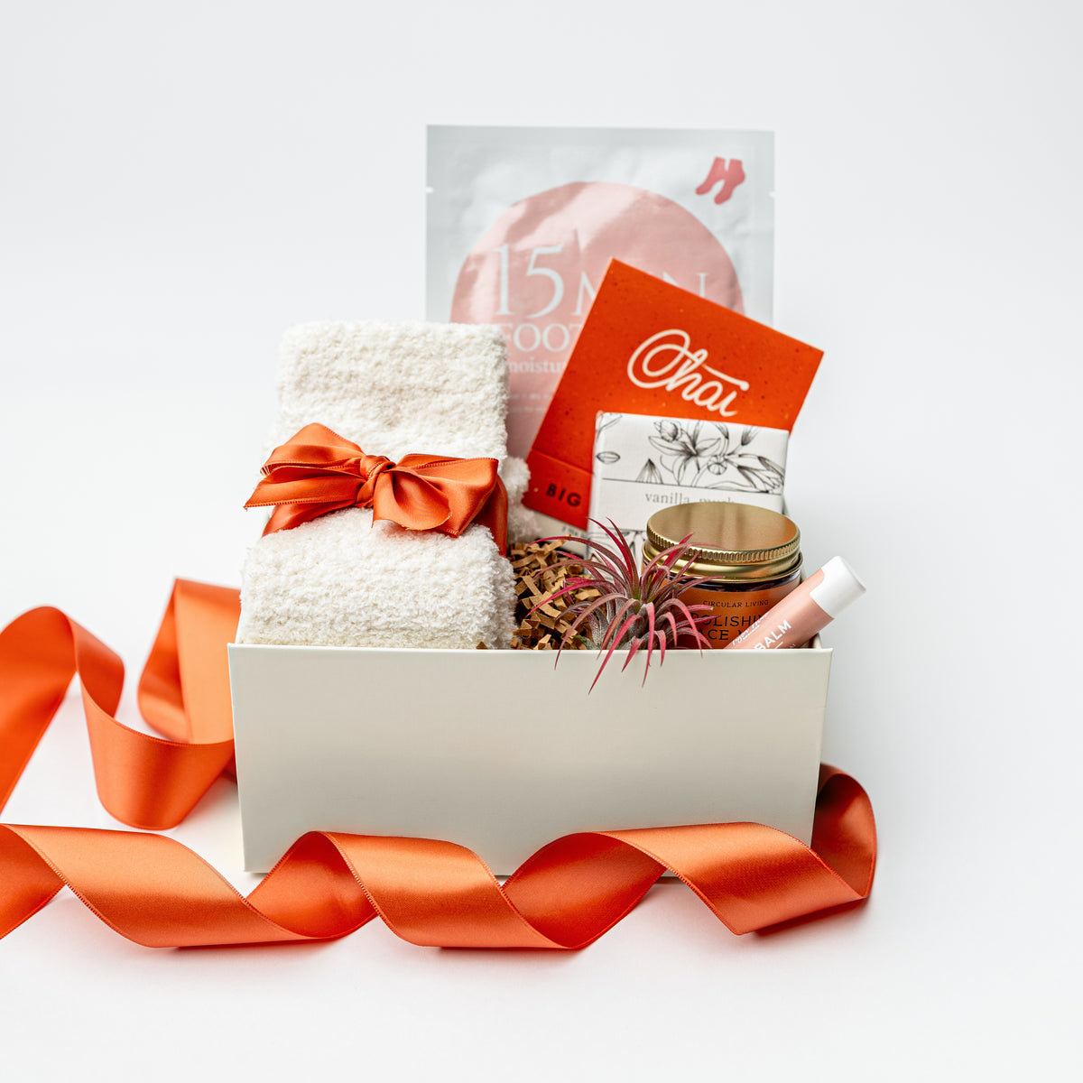 A curated gift box set on a white background, featuring a white textured towel tied with an orange satin ribbon, a pink facial mask packet, an orange card labeled 'Choi', a small jar with a gold lid, and a lip balm tube.