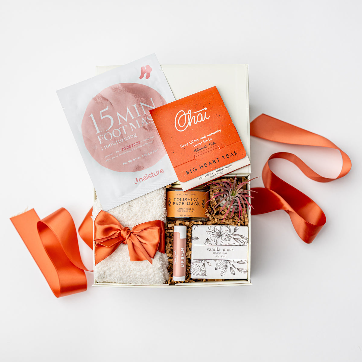 A curated wellness gift box set on a white background, featuring a 15-minute foot mask packet, a tea packet, a jar of foot mask powder, a lip balm, and a boxed item tied with an orange satin ribbon.