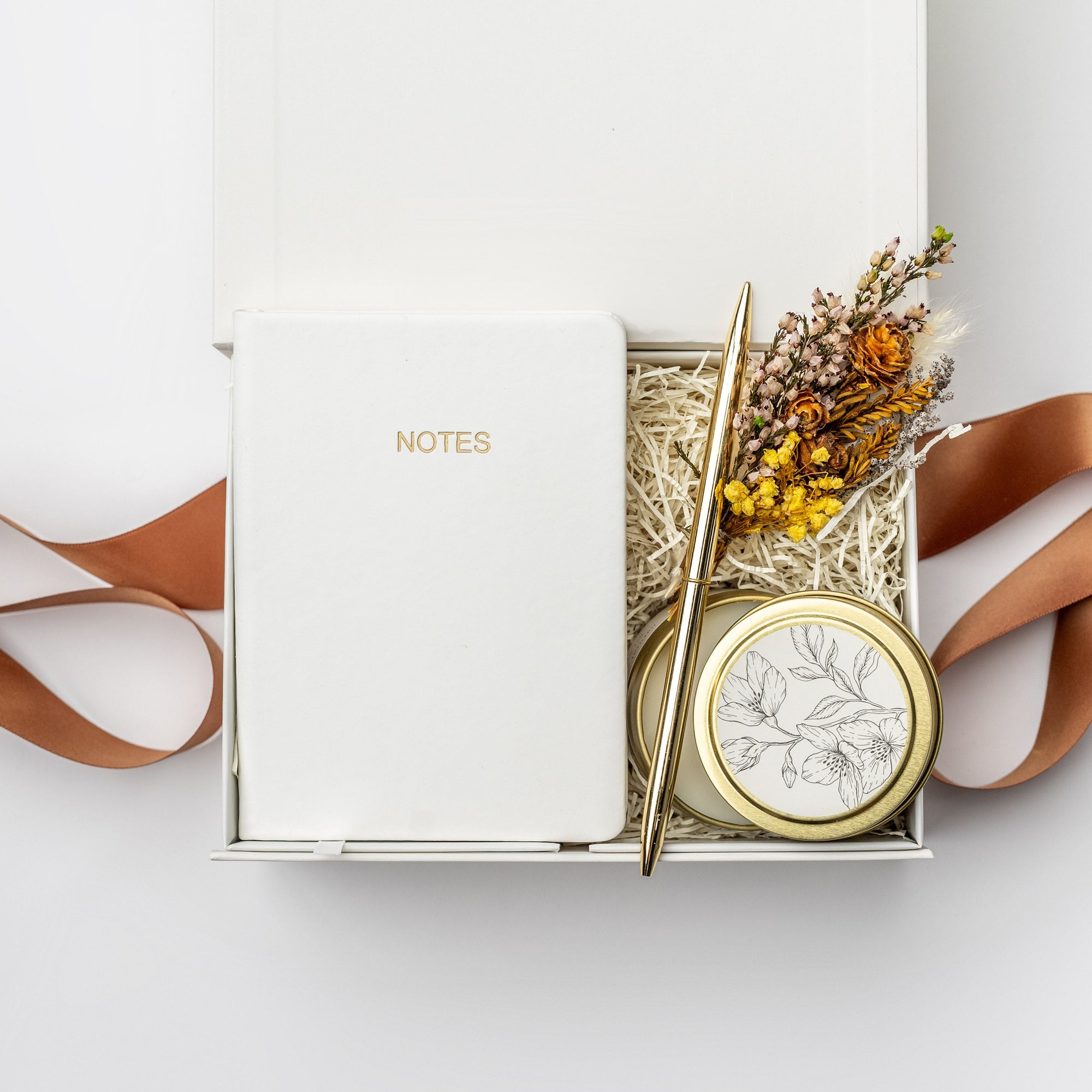 A top view of the Thankful For You Gift Box presented in a white gift box filled with ivory shred and tied with a toffee-colored ribbon, featuring an ivory softcover pocket journal, a minimalist gold bullet pen, a 4 oz. hand-poured White Tea candle, and a handmade dried floral mini bouquet, all arranged on a white background.