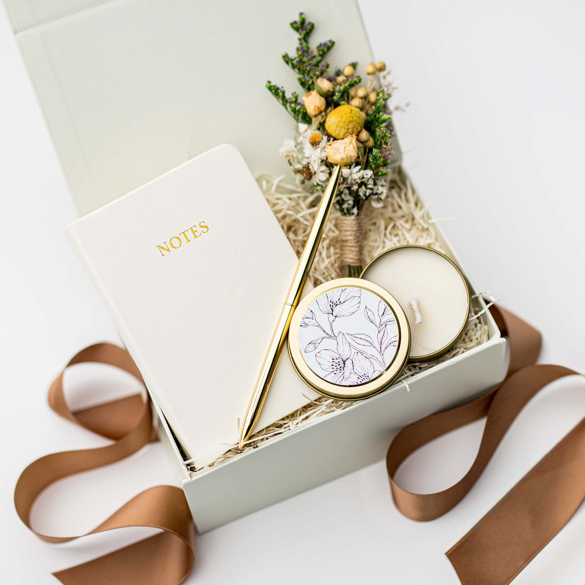 A top view of the Thankful For You Gift Box presented in a white gift box filled with ivory shred and tied with a toffee-colored ribbon, featuring an ivory softcover pocket journal, a minimalist gold bullet pen, a 4 oz. hand-poured White Tea candle, and a handmade dried floral mini bouquet, all arranged on a white background.