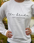 The bride custom name sweatshirt in white personalized in black lettering the bride Mrs. Anderson worn by the bride ina nature background. 
