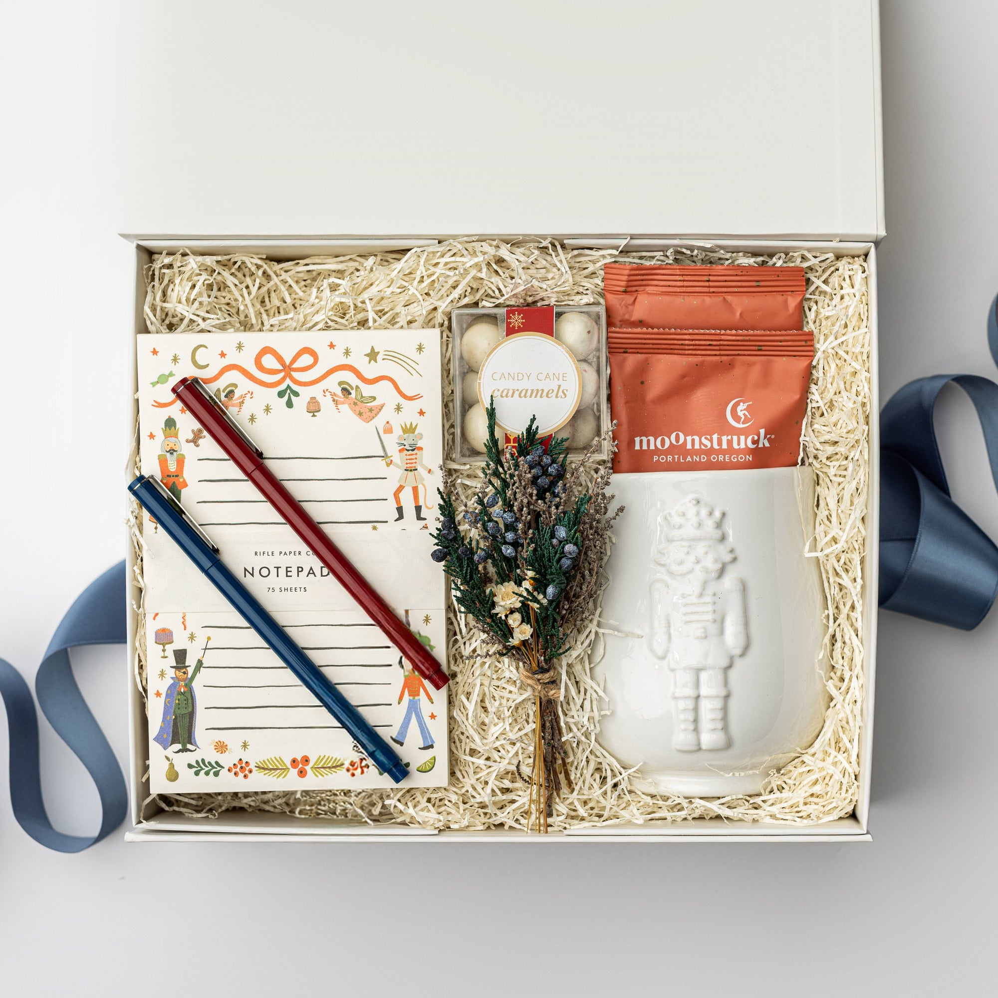 The Nutcracker Holiday Gift Box is featured open, revealing a white gift box with a blue ribbon crossing underneath. Inside the box, all arranged on top of white shred. On the left side, there is a Nutcracker notepad with a blue and red pen on top. In the center, a clear box of candy caramels with a white label, gold rim, and red ribbon sits beside a floral bundle. On the right, there is a white embossed Nutcracker mug containing two orange monstruck chocolate packages, all on a white background.