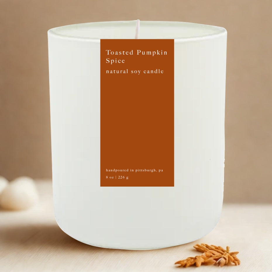 Toasted pumpkin spice candle featured in its white ceramic jar with orange label with white lettering on a beige background with fall details. 