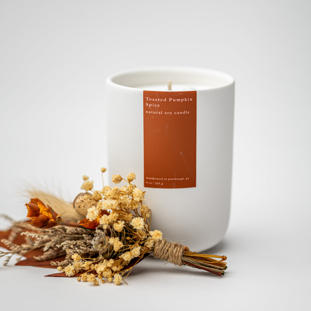 A white soy candle jar with a terracotta orange label reading 'Toasted Pumpkin Spice natural soy candle' on a light gray background, styled with dried flowers and grasses in front.