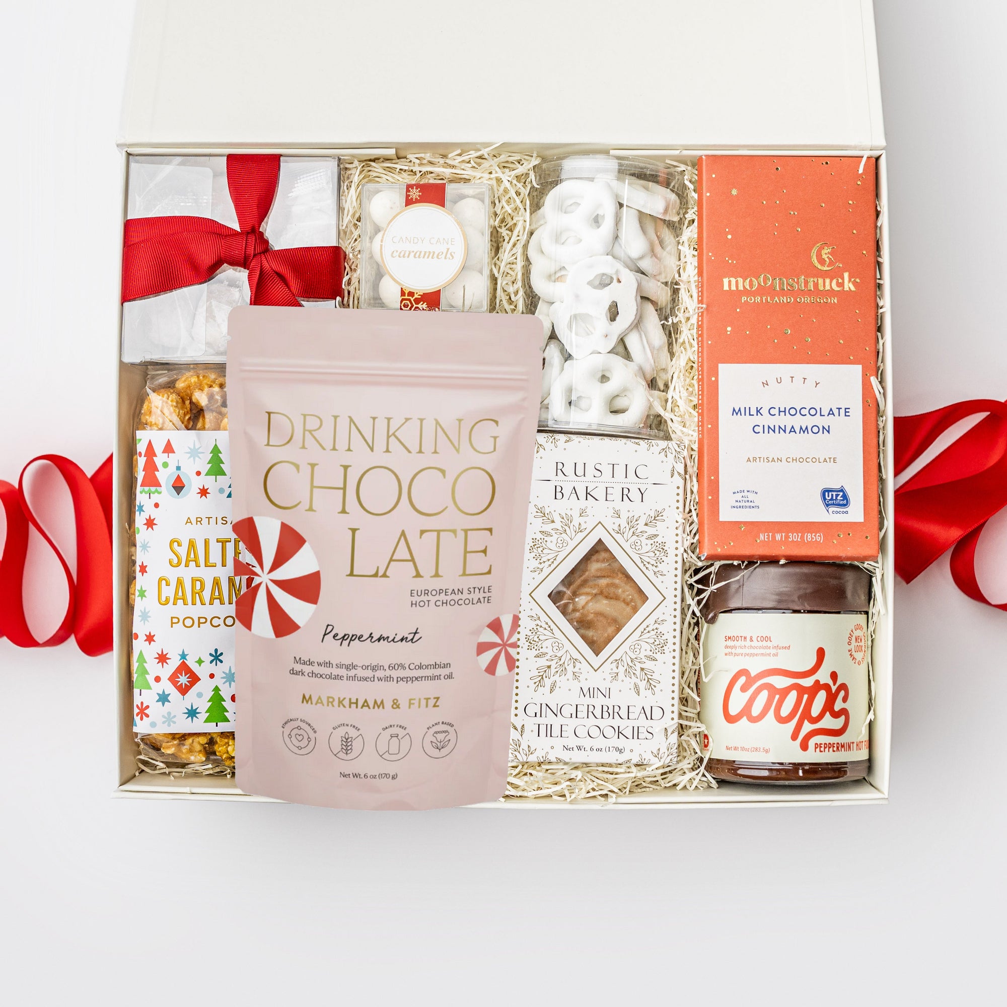 A white gift box with ivory paper shred and a red ribbon on a white background. Inside the box are Markham & Fitz peppermint drinking chocolate, Lark artisan tea cookies, rustic bakery gingerbread tile cookies, white chocolate pretzels, Art of Caramel salted caramel popcorn, candy cane caramels, candy cane striped gummies, and Coops peppermint hot fudge — a festive “Treats for All” holiday gift box.