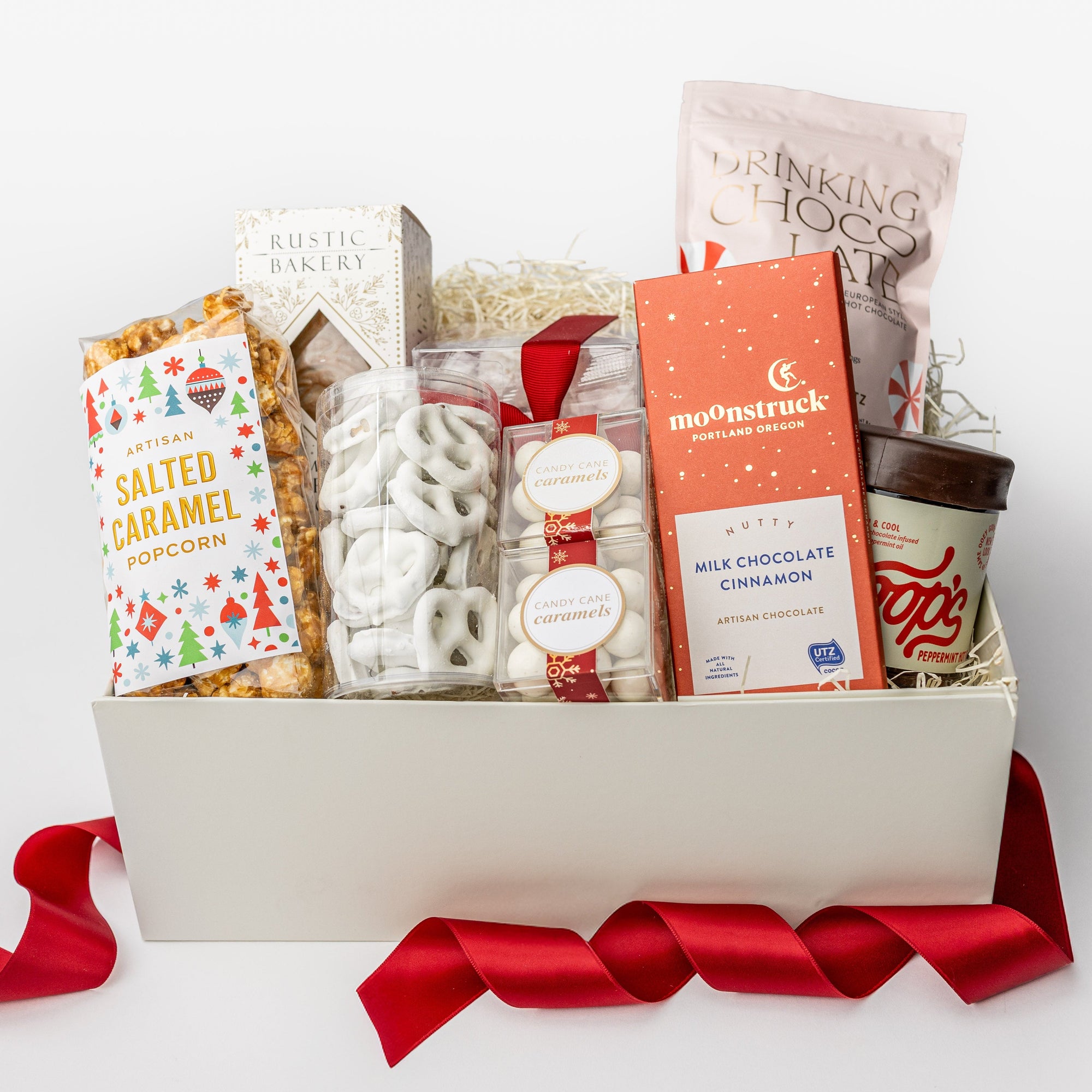 A white gift box with ivory paper shred and a red ribbon on a white background. Inside the box are Markham & Fitz peppermint drinking chocolate, Lark artisan tea cookies, rustic bakery gingerbread tile cookies, white chocolate pretzels, Art of Caramel salted caramel popcorn, candy cane caramels, candy cane striped gummies, and Coops peppermint hot fudge — a festive “Treats for All” holiday gift box.