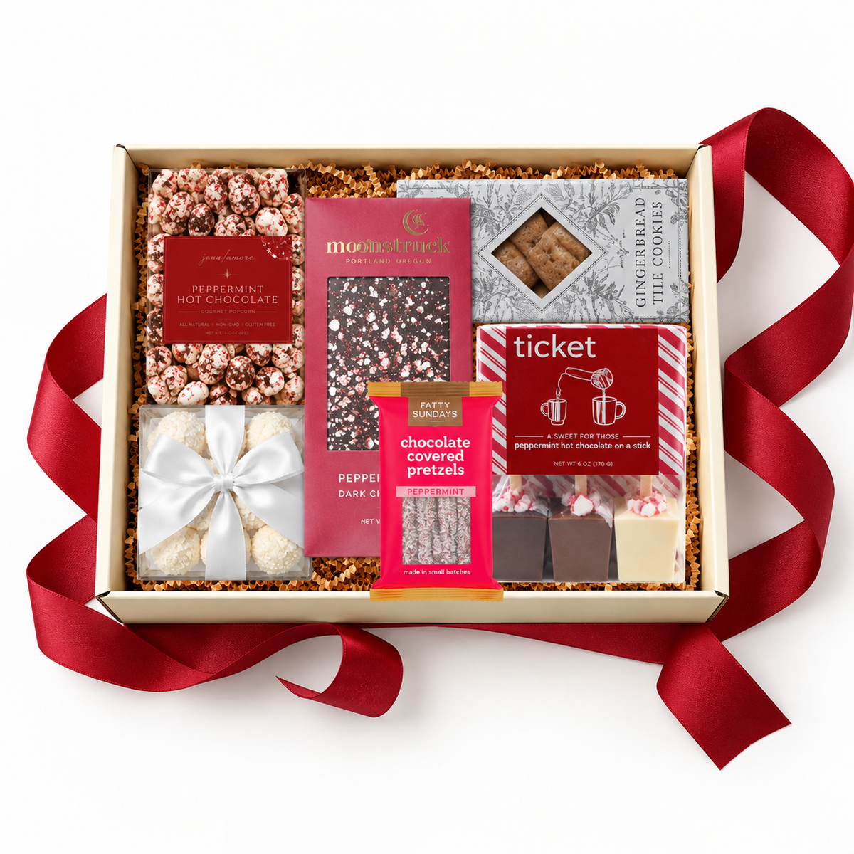 A gourmet chocolate and snack gift box arranged in a beige box with shredded paper filler and a red ribbon. Visible items include peppermint hot chocolate, peppermint-covered pretzels, dark chocolate, gingerbread cookies, and candy, with several packages showing red and pink branding.