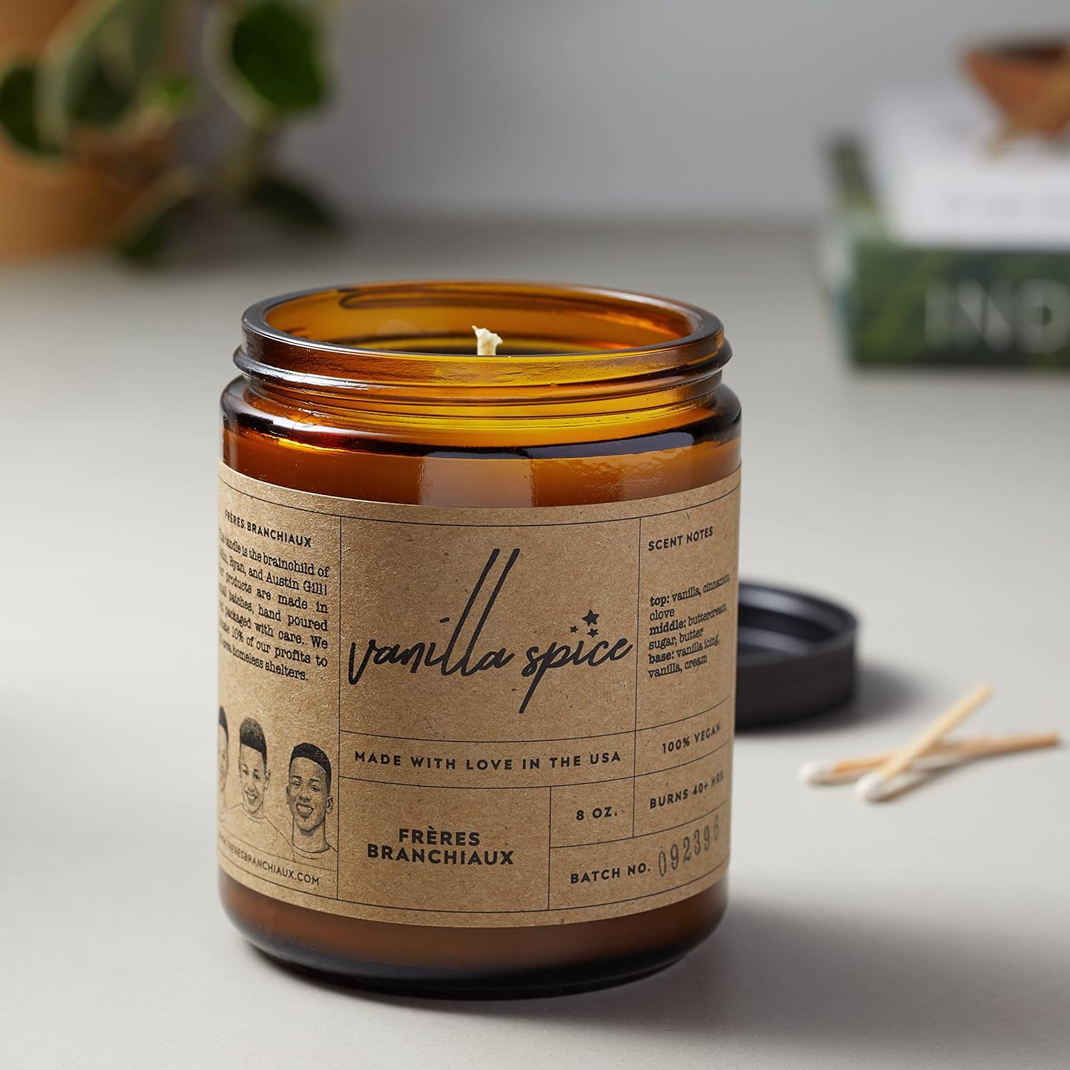 Vanilla Spice Candle form Frères Branchiaux, showcasing the candle in its brown jar and paper-craft light brown label with black lettering opened.  In a white background with the black lid of the candle, white tip matches and books and plants blurred in. 