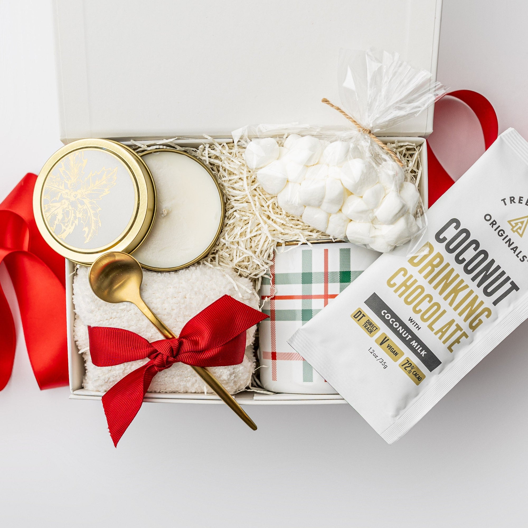 A white gift box filled with ivory paper shred, accented with a red ribbon around and under a pair of plush ivory cloud socks, on a white background. Inside the box are a 12 oz holiday plaid gold‑rimmed ceramic mug, a single‑serve packet of Treehouse Originals coconut hot cocoa, mini marshmallows, a 4 oz hand‑poured candle labeled “‘Tis the Season,” and a gold stirring spoon — a cozy holiday gift set.